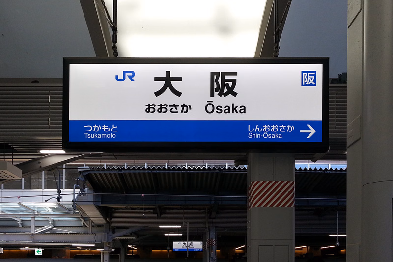 A large electronic sign at a train station displays "JR 大阪" and "おおさか Osaka", with directions to "つくもと Tsukamoto" and "しんおおさか Shin-Osaka".