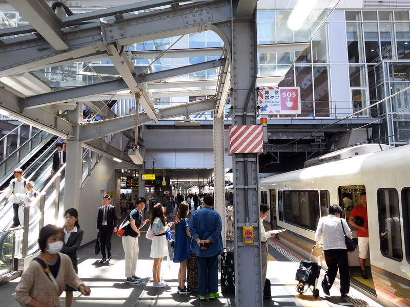 A bustling train station in Ōsaka, Japan, with people waiting for their train and signage indicating the station's name and direction.