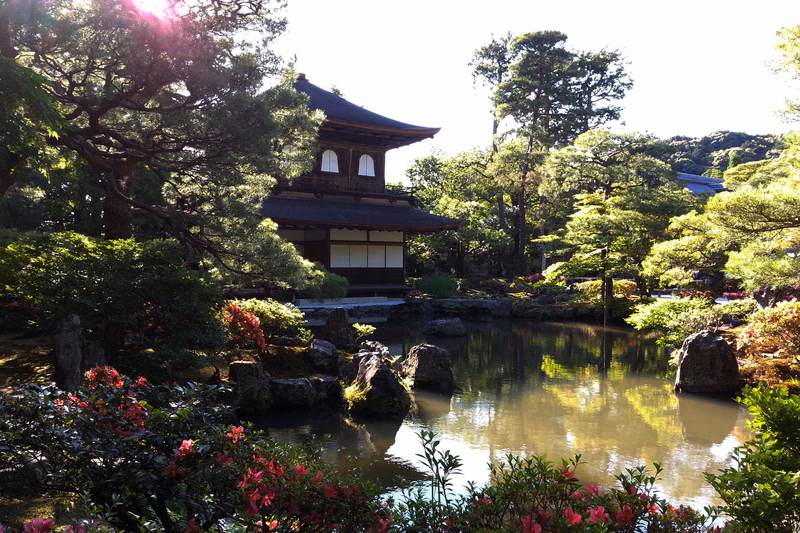 A traditional Japanese temple building is nestled beside a calm pond, surrounded by lush green trees and colorful flowers.
