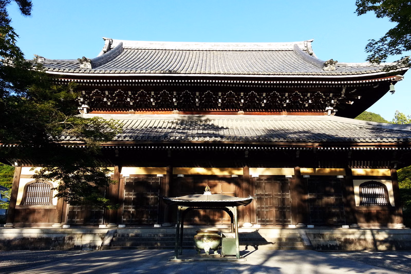 A traditional Japanese temple building with a tiled roof stands in a courtyard featuring a stone lantern in the foreground.