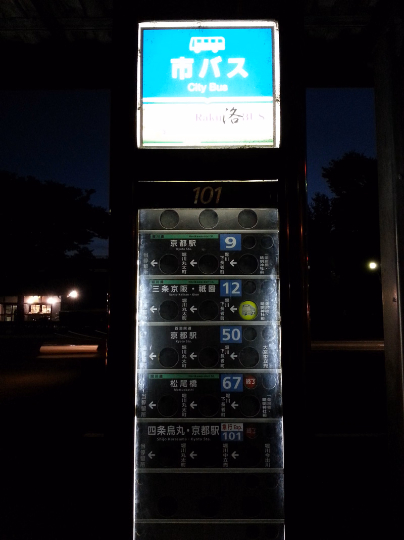 A vertical bus stop sign displays "市バス City Bus" and lists route numbers like 101, 9, 12, 50, and 67.