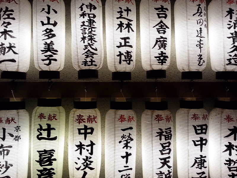 A row of illuminated Japanese paper lanterns displaying vertical text, including characters like "古舎廣幸" and "中村波".