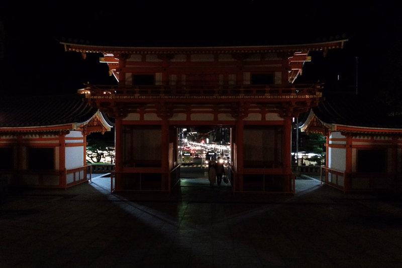 A large, red wooden torii gate stands at the entrance of a temple complex at night, with lights visible through the doorway.