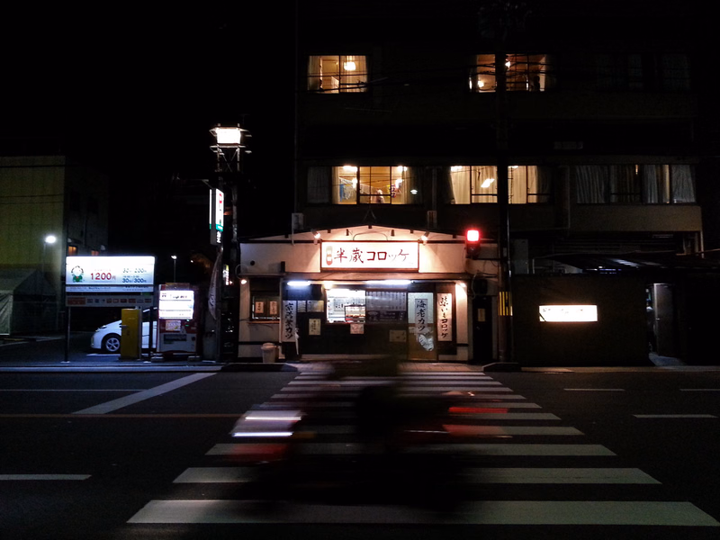 A nighttime street scene featuring a restaurant with a sign that reads "半世紀コロワ" and a crosswalk in the foreground.