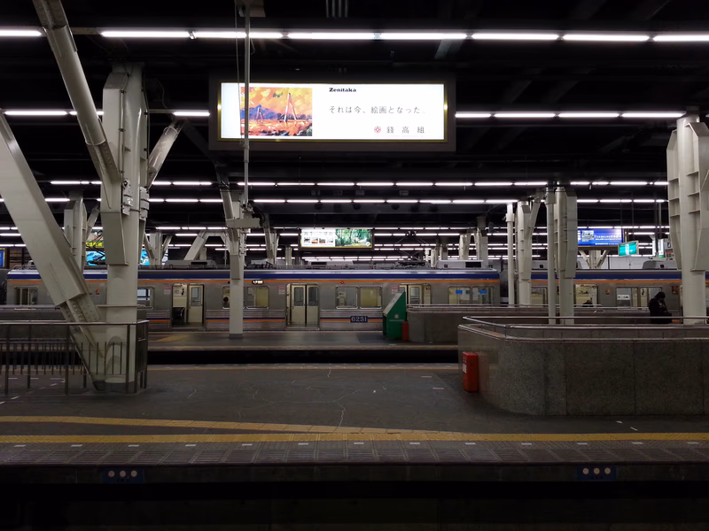 A photograph of a train station in Osaka, Japan. The image shows a modern train station with a sleek design, featuring a large digital display above the platform and a few passengers waiting for the train.