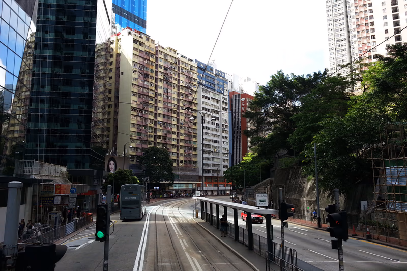 A photo of a city street with a tram, a bus, and tall buildings, with trees and a traffic light in the foreground.