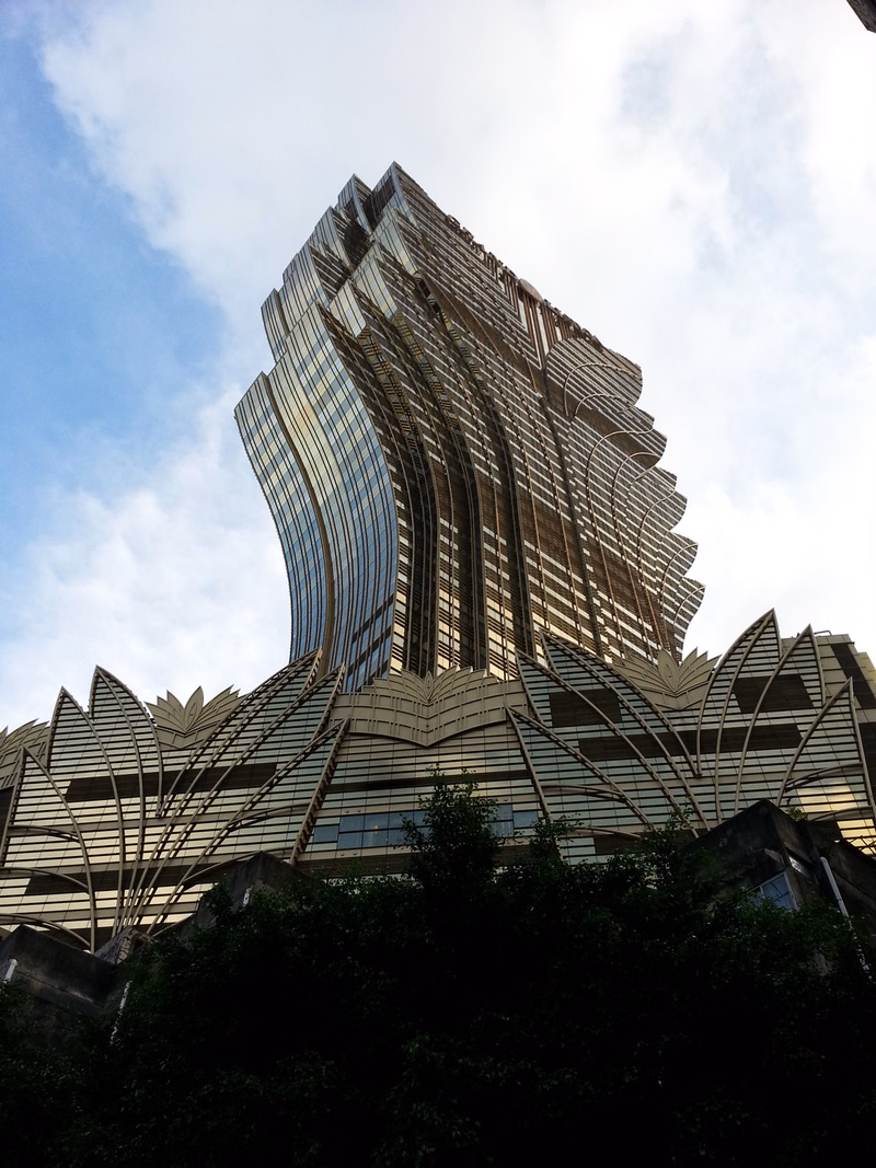 A modern, textured skyscraper with a complex, layered facade rises against a bright, cloudy sky.