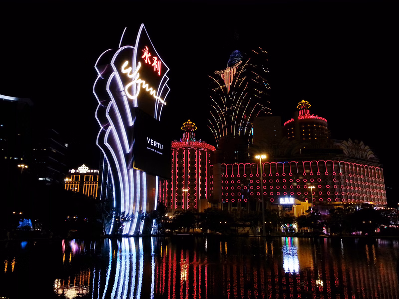 A nighttime view of a large casino complex with bright illuminated signs, including one displaying "VERTU" and Chinese characters, reflected in the water.