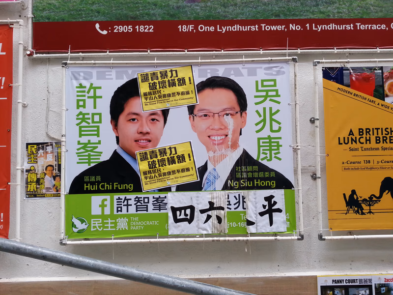A poster advertising a political campaign in Hong Kong. The poster features two men, one in a suit and the other in a traditional Chinese outfit, with Chinese text and English text. The English text includes the name 'Hui Chi Fung' and 'Ng Siu Hong.' The Chinese text is a political advertisement.