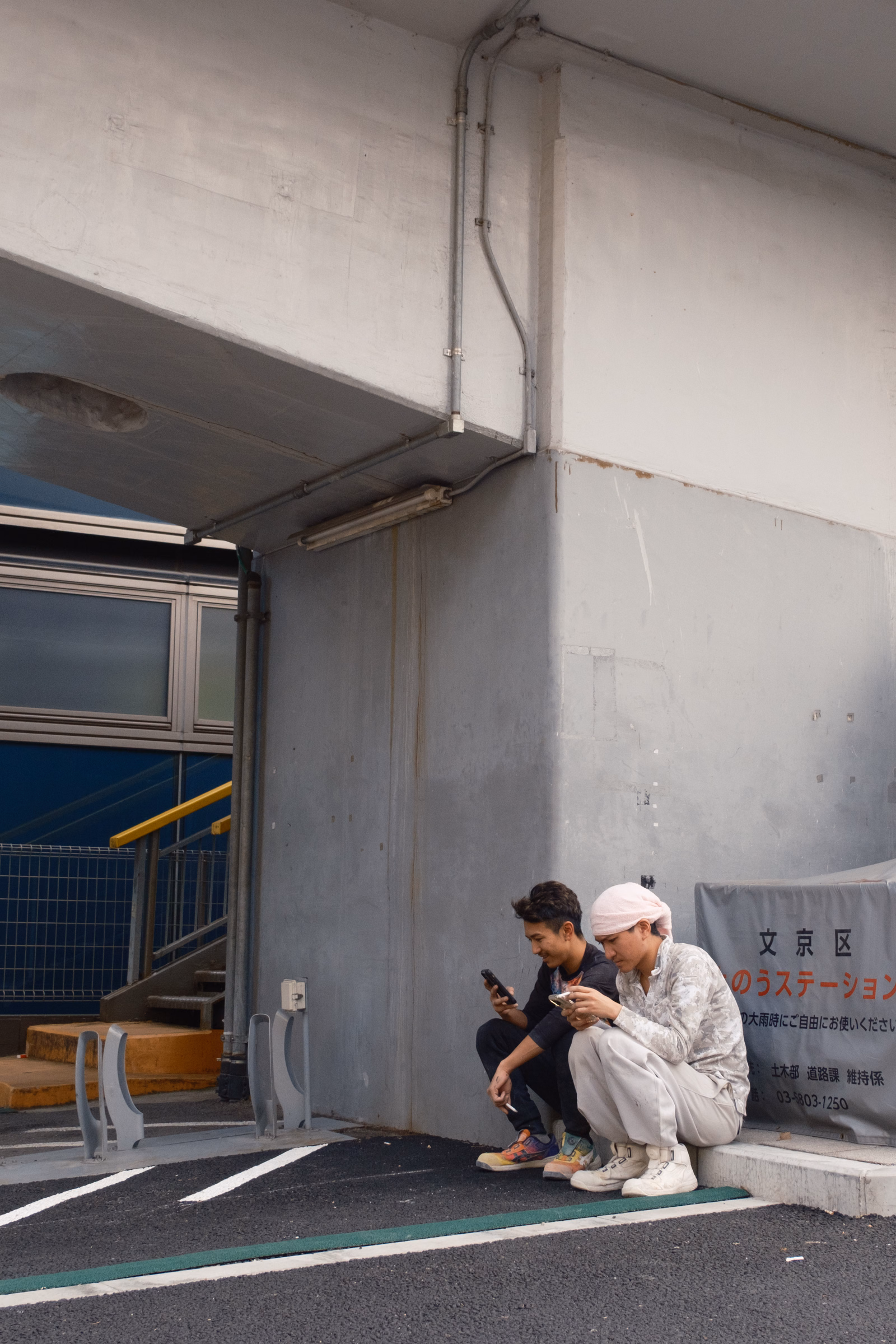 Three people sit near a concrete wall outside a station, with a sign that reads "文京区 のうステーション".