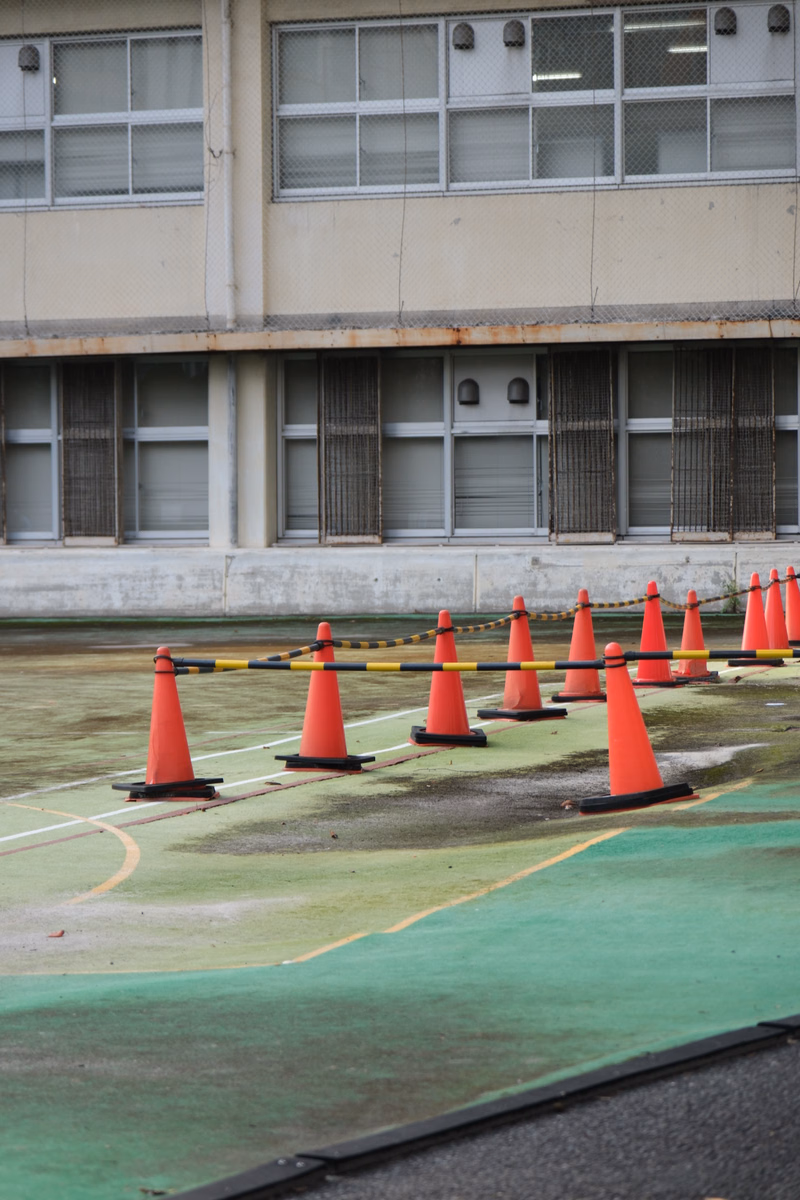 A line of orange traffic cones is set up on a grassy sports field in front of a large, light-colored building.