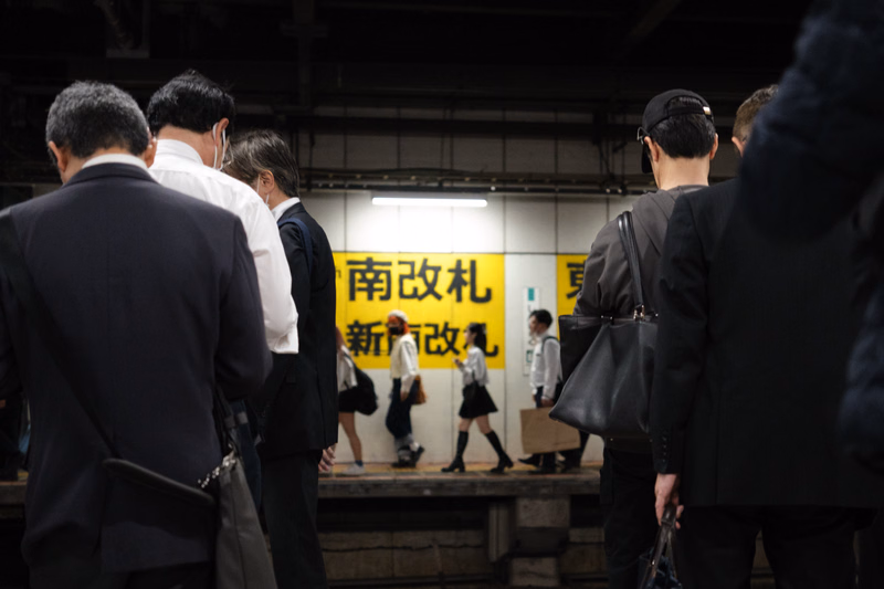A group of businessmen in suits stand indoors in front of a yellow sign that reads "南改札 新南改札".