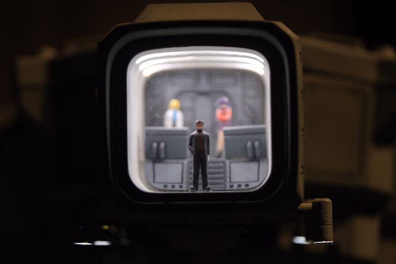 A small action figure stands inside a clear, square display case, with other miniature figures visible behind it.