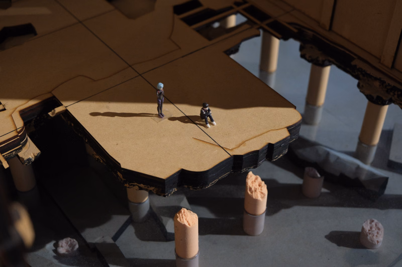 Two small miniature figures stand on a large, brown, cut-out piece of cardboard representing a building section.