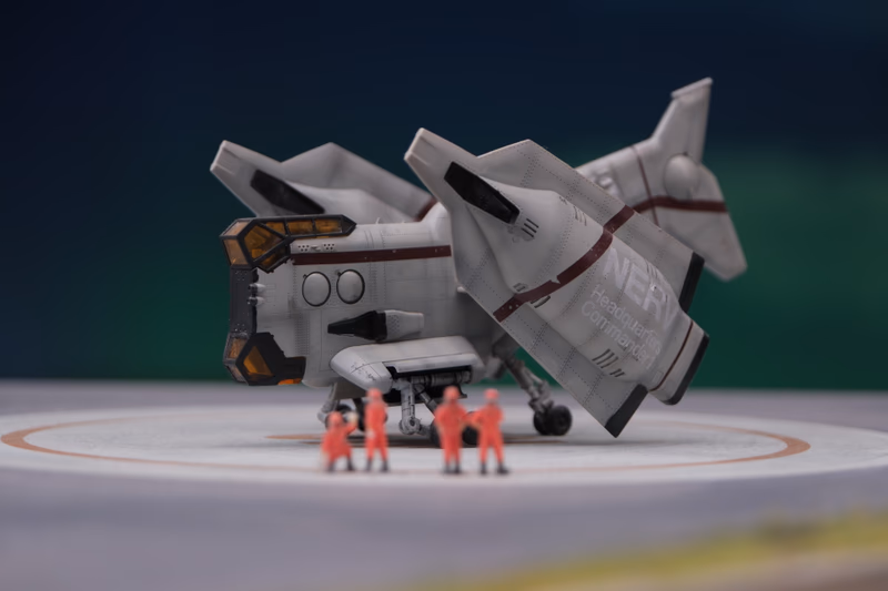 A futuristic spacecraft with the text 'NER' and 'GUNS' visible on its side. The spacecraft is situated on a circular platform, and there are small figurines of people in red near the bottom of the image. The background is blurred, focusing on the spacecraft and the people.