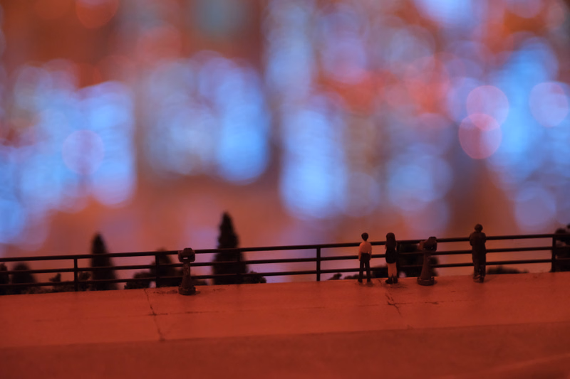 Several small figures stand along a railing overlooking a blurred, colorful cityscape during sunset.
