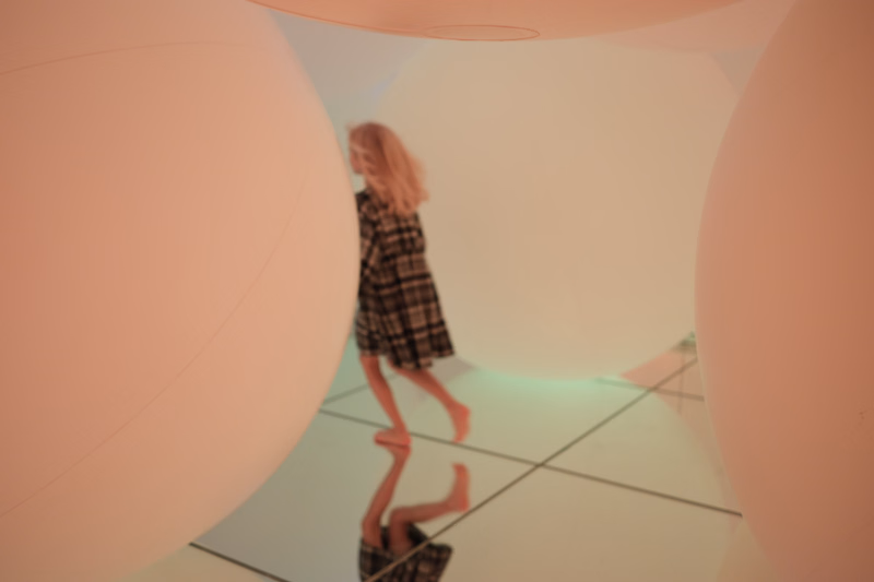 A woman in a plaid dress stands inside a minimalist room surrounded by large, smooth, peach-colored spheres.