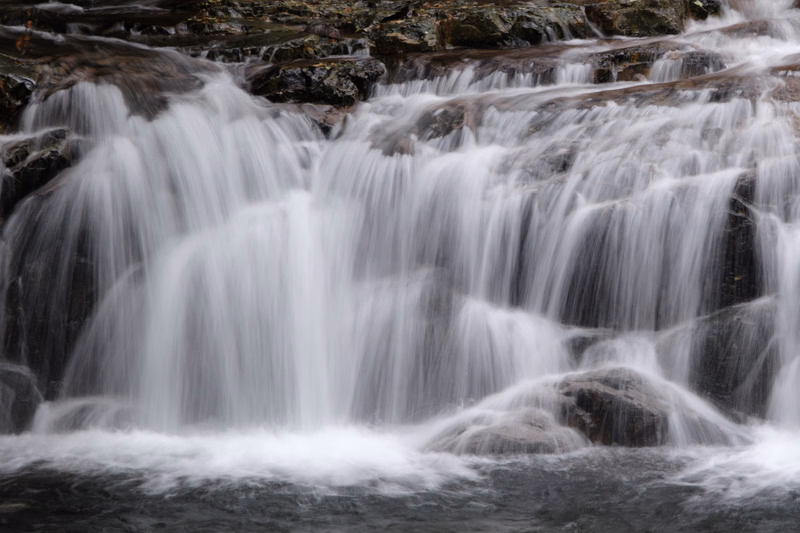 A wide shot captures a multi-tiered waterfall cascading over dark, wet rocks into a pool below.