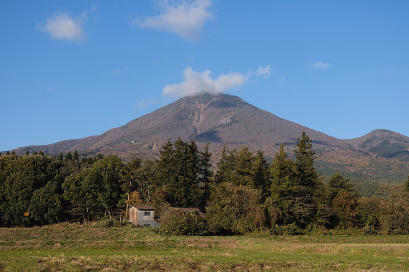 A large, conical mountain or volcano rises above a dense forest under a bright blue sky with scattered white clouds.