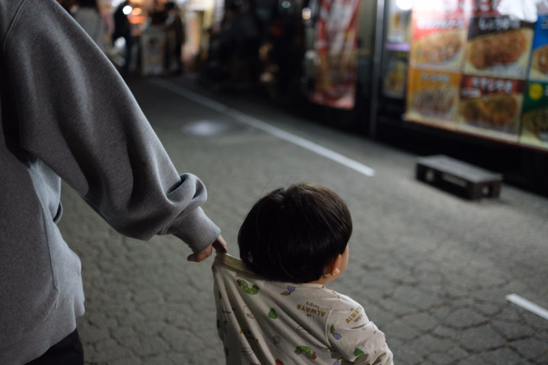A small child looks at a food stall while an adult leans down toward the child on a paved street.