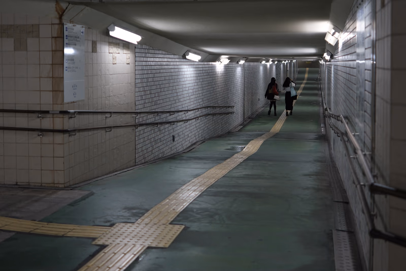 A dimly lit underground tunnel with a few people walking down the center, surrounded by tiled walls and metal railings. The floor is a mix of green and yellow tactile paving.
