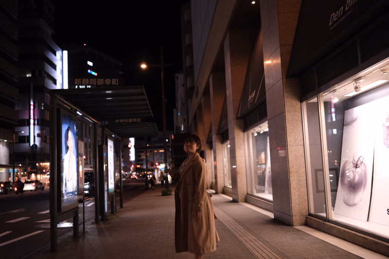A woman in a long coat walks down a city sidewalk at night, past illuminated storefronts.