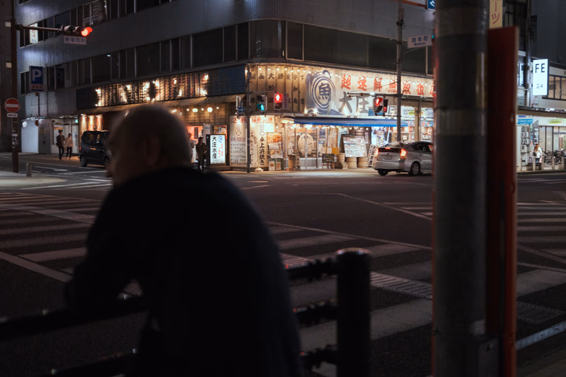 A man sitting on a bench in front of a brightly lit building at night.
