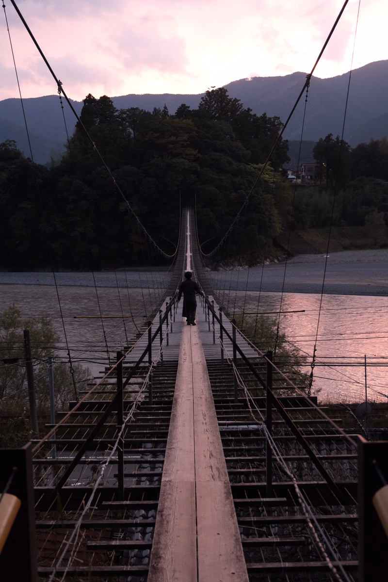 A lone person walks across a long suspension bridge over a river during a pink and purple sunset with mountains in the background.