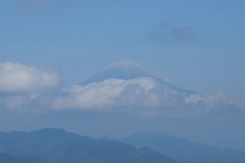 A large, conical mountain or volcano is partially obscured by a layer of white clouds under a clear blue sky.