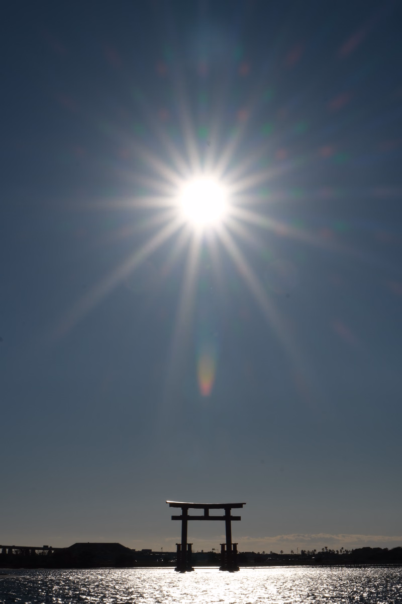 A silhouette of a torii gate stands against a bright sun setting over the water.