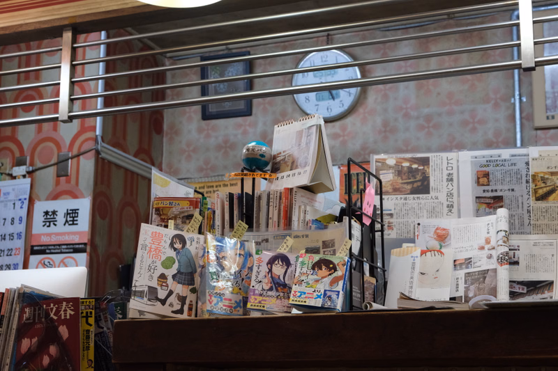 A cluttered shelf in a shop displays various magazines and books, including one with the text "禁煙" and another with "No Smoking".