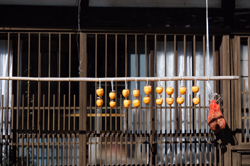 Several skewers of grilled meat and small yellow food items are hanging from a bamboo rod outside a wooden building.
