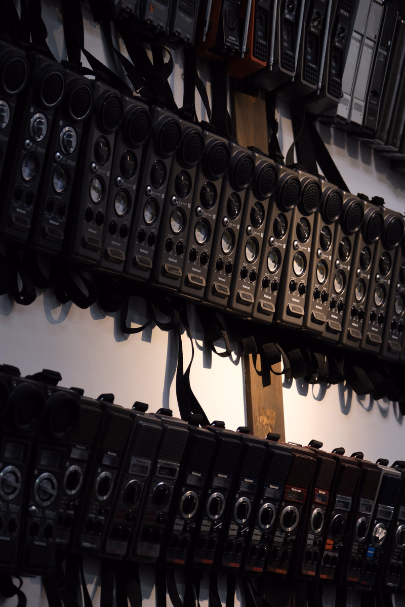 A row of professional audio mixing consoles and soundboards are stacked on shelves in a studio setting.