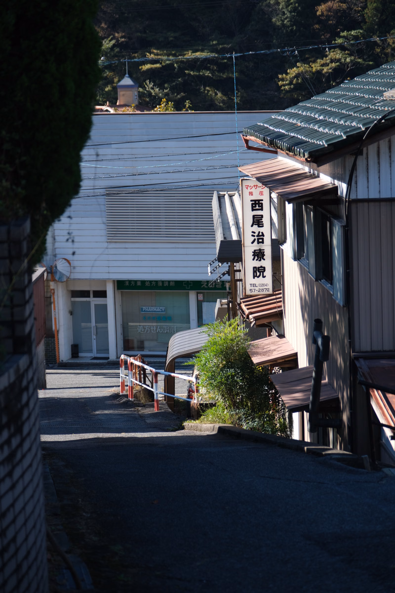 A street view showing a white building with a pharmacy sign that reads "西尾治療院" and a smaller storefront next to it.