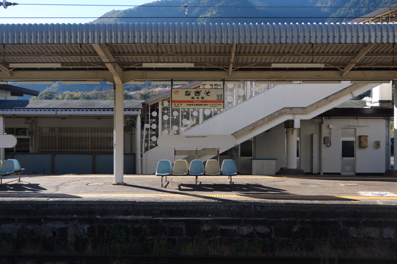 A covered train station platform features a sign that reads "なぎそ" and several blue benches under the shelter.