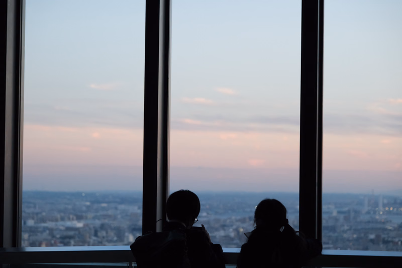 Silhouettes of three people look out of a large window at a sprawling city view during sunset.