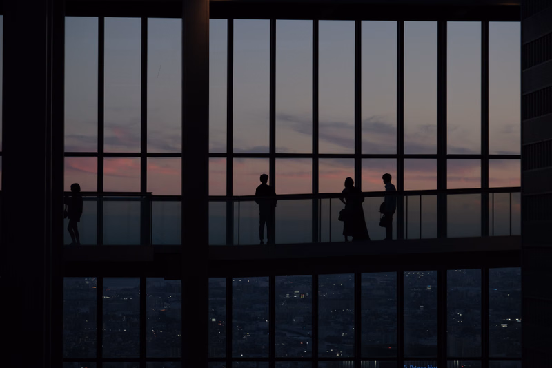 Silhouettes of several people stand on a balcony overlooking a city skyline during a colorful sunset.