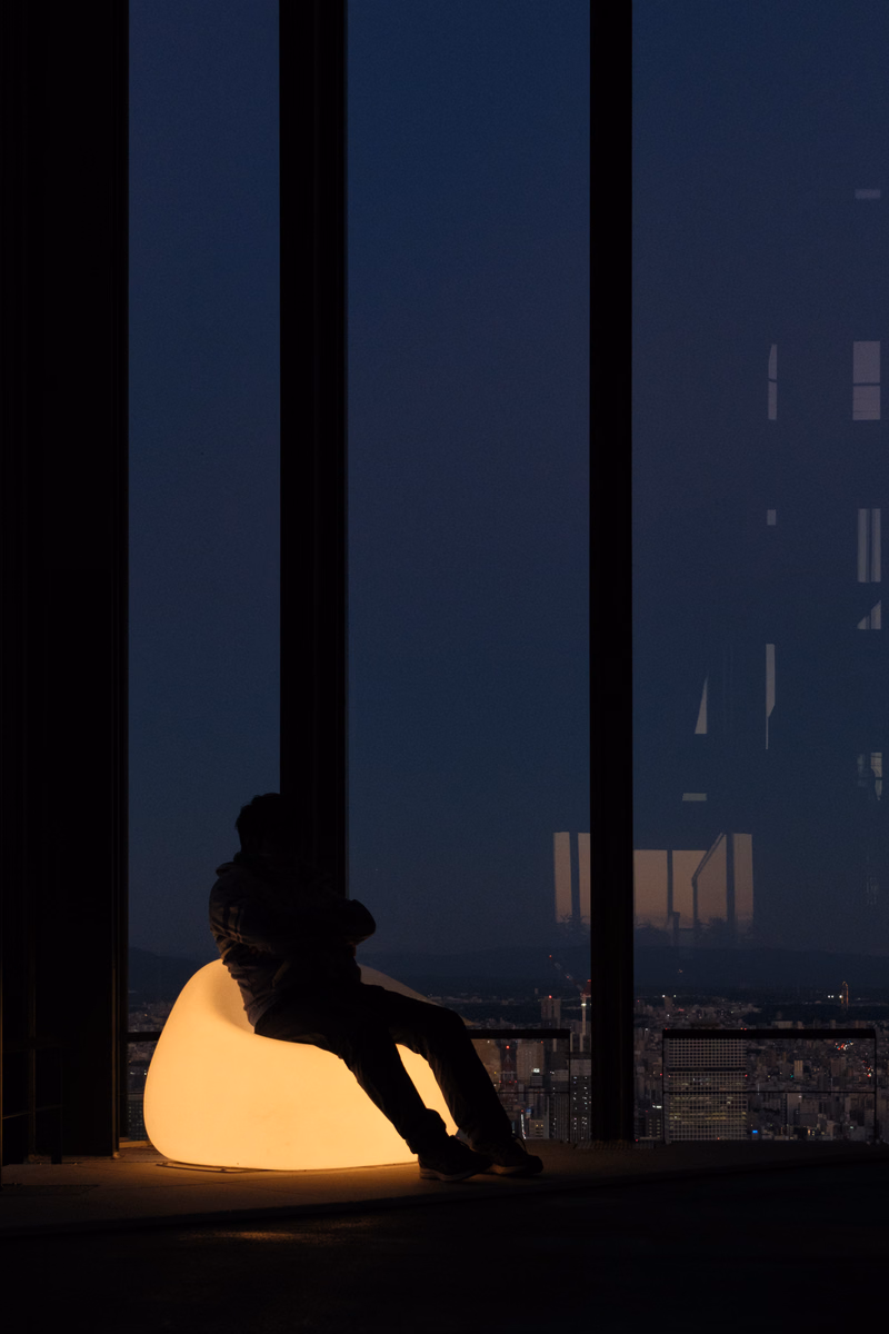 A silhouette of a person sitting in a glowing chair looking out a large window at a nighttime city view.