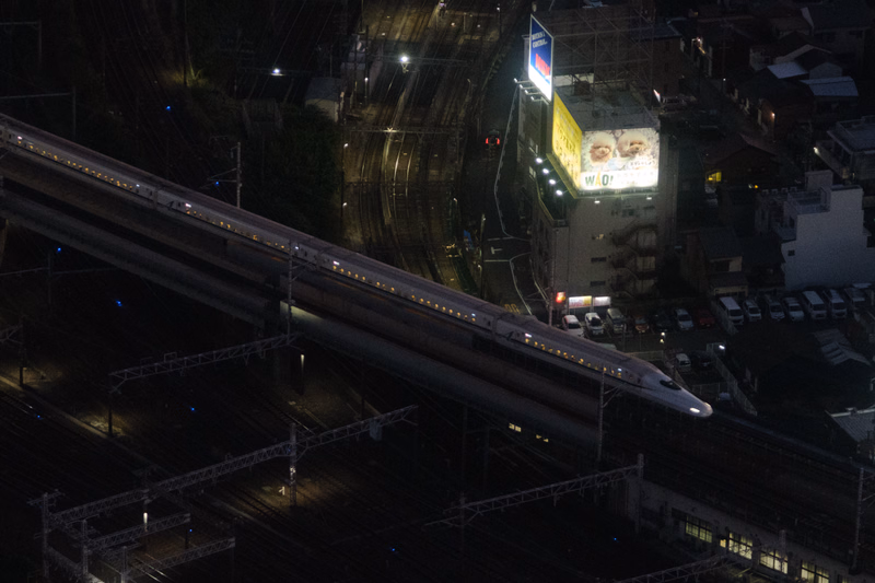 A high-speed train travels along an elevated track near a brightly lit building displaying the text "WAO!" at night.