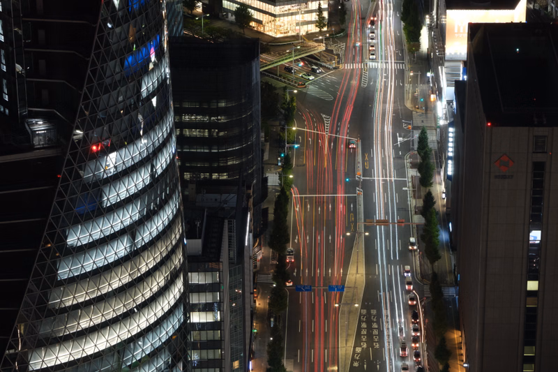 A nighttime cityscape view showing busy traffic flowing through a multi-lane city street lined with modern buildings.