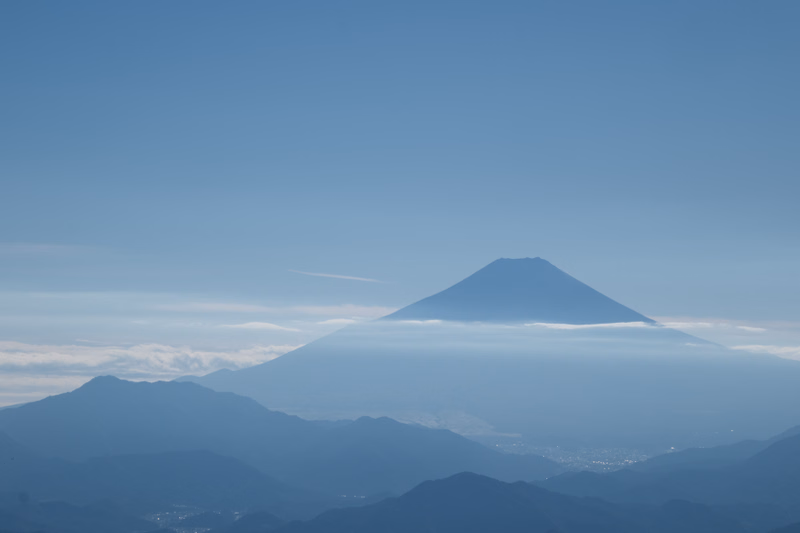 A large, conical mountain peak rises above a layer of low clouds and haze under a clear blue sky.