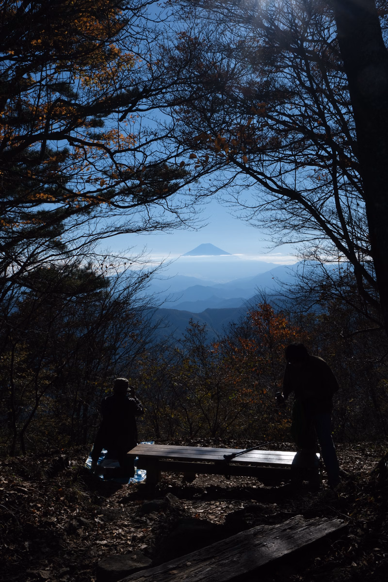 Silhouettes of people sit near a wooden bench overlooking a vast mountain landscape with a prominent peak in the distance.