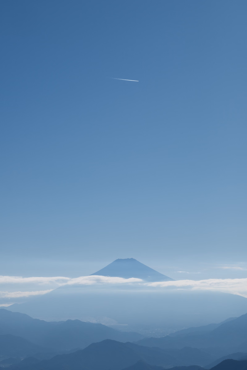 A large mountain peak rises above a layer of clouds under a clear blue sky, with a faint airplane contrail visible.