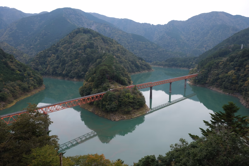 A red bridge crosses a serene lake surrounded by lush green mountains and forests.