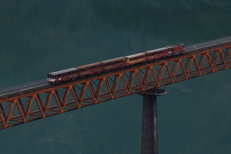 A passenger train crosses a large, orange truss bridge over a body of dark green water.