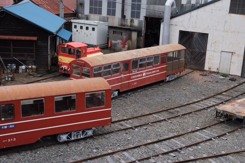 Several vintage red and white train cars, including one labeled "OIGAW RAILWAYS", sit on railway tracks near various industrial buildings.