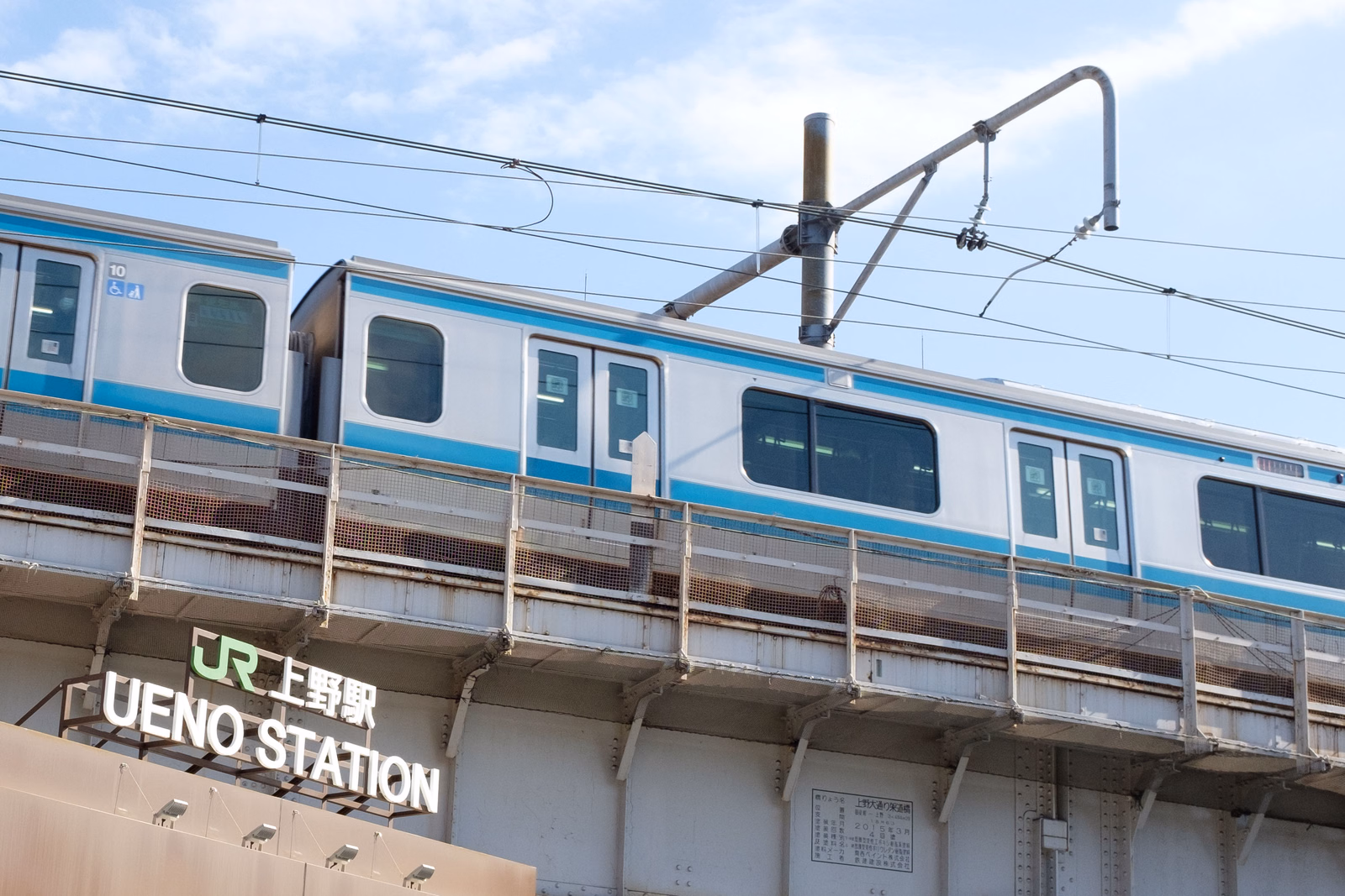 A blue and white train is stopped at Ueno Station, with a sign reading "JR UENO STATION" visible below the tracks.