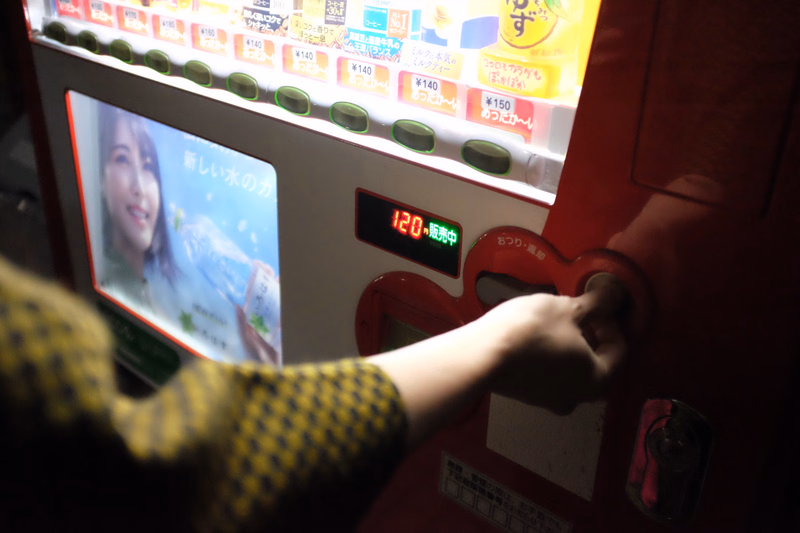 A person's hand is inserting money into a red vending machine that displays "120・販売中" and features a screen with a woman's image.