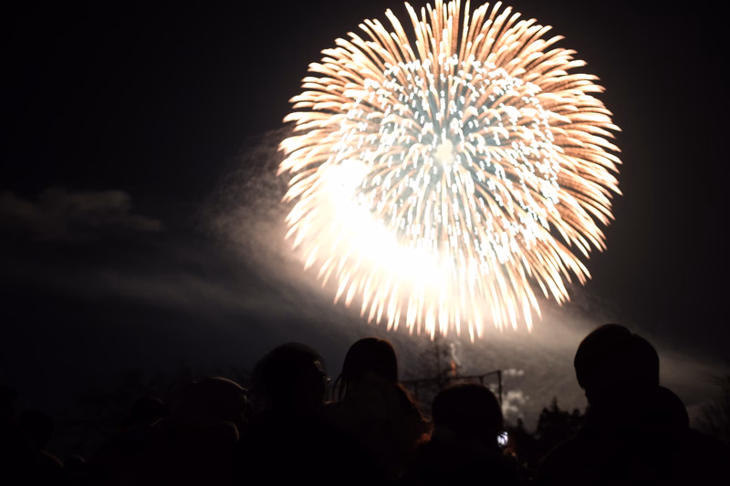 A large, bright fireworks display illuminates the dark night sky above a silhouetted crowd of people.