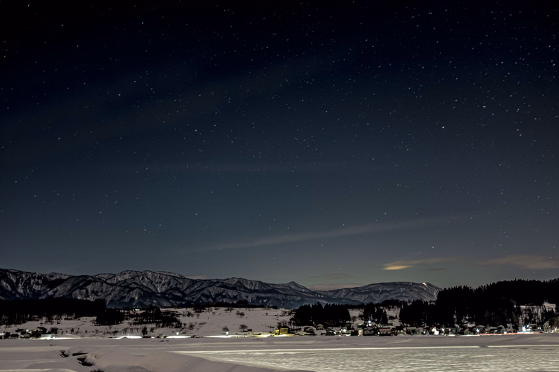 A wide-angle photograph of a starry night sky over snow-covered mountains and a frozen body of water.